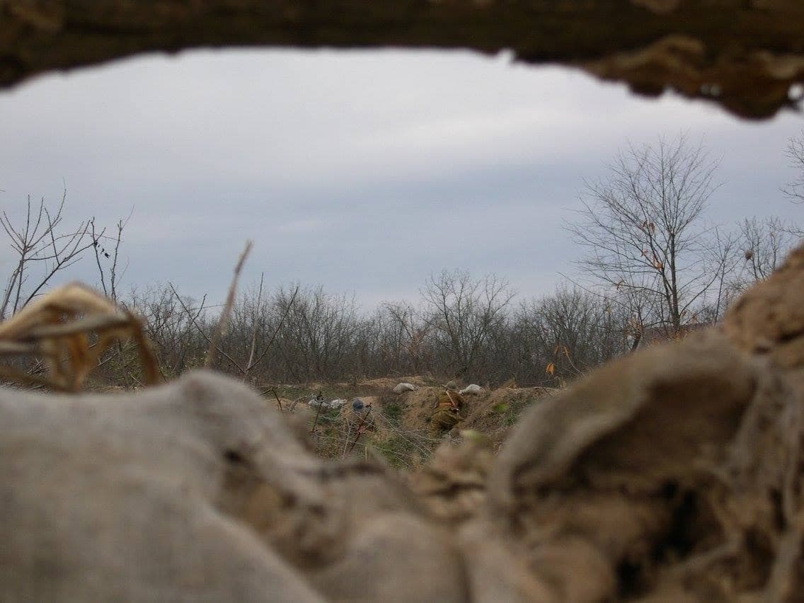 Modern color photograph shot through a loophole in a sandbagged wall, looking out over a bare woodland with a reenactor visible in a shell hole in the distance.