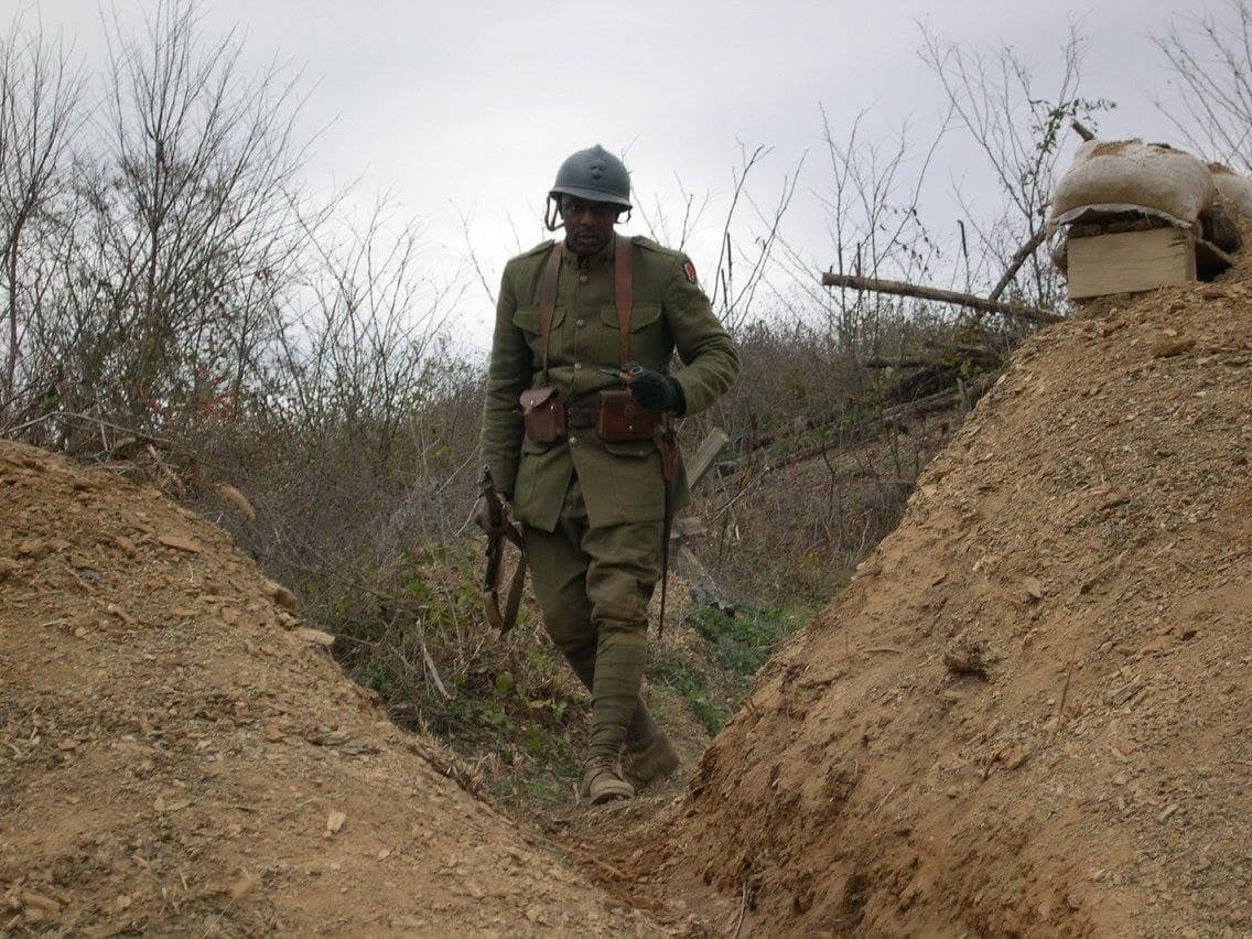 Modern color photograph of a reenactor walking through a dug trench, rifle with fixed bayonet slung over his shoulder.