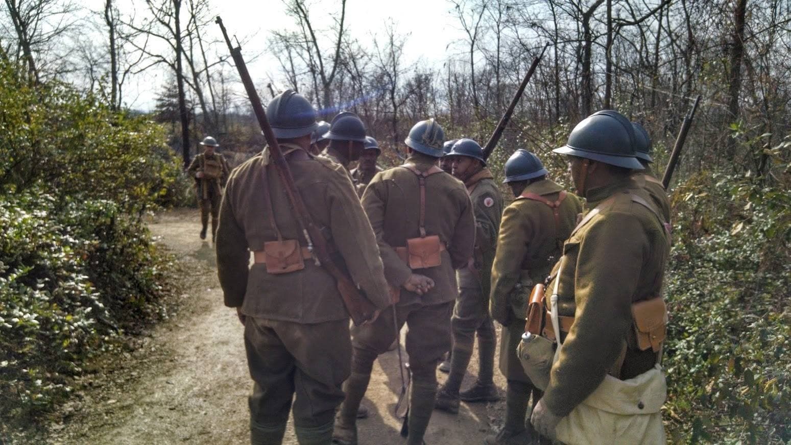 Modern color photograph, viewed from behind, of reenactors walking a wooded trail with rifles slung and first-aid pouches visible on their belts.