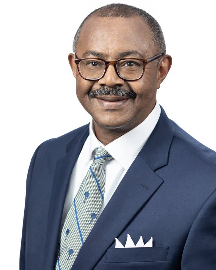 Studio portrait of Perry James: an older Black man with a grey moustache and tortoiseshell glasses, smiling, wearing a navy suit, white shirt, and a light green tie patterned with small tree motifs, photographed against a plain white background.